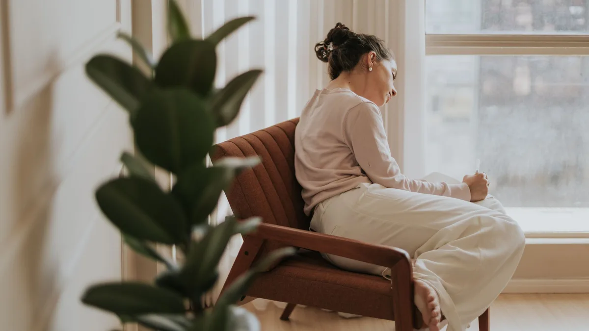 A woman sitting by a large window with a plant, thinking through questions about GoHighLevel plans