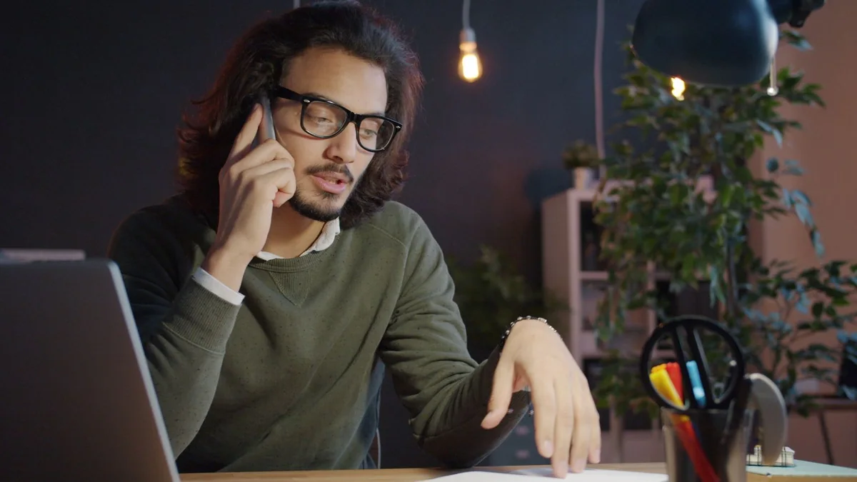 A man with glasses talks on his phone beside an open laptop, from our GoHighLevel support guide